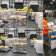 Warehouse lumpers stacking boxes from a freight truck into a pallet, wearing safety vests and gloves inside a busy distribution center